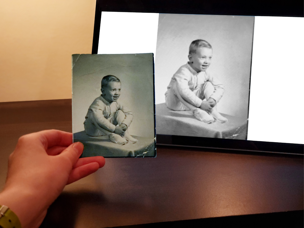 A hand holding a photograph in front of a computer screen displaying the same photograph